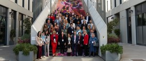 Various diversely clad experts and professionals stand for a group picture outdoors, at the foot of a grey, modern staircase, located between two offices 