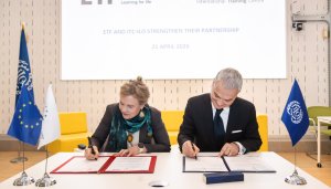 On a white table, a blonde agency director with a grey blazer and green scarf signs an agreement alongside a grey-haired gentleman wearing a suit