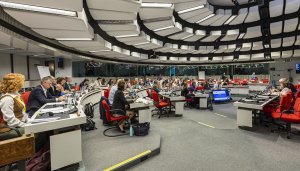 A brightly lit, largely white and grey hemicircular conference room, viewed in side profile, filled with people sitting down and observing the centre