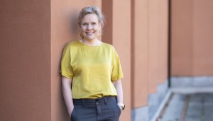 Pilvi Torsti, wearing a yellow satin top and grey slacks, smiling and leaning against the terracotta-coloured faced of a colonnade-ripple building