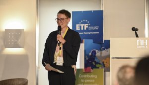 A short-haired young woman, piece of white paper in hand, wearing glasses, a grey jacket and a lanyard, holding a microphone as she addresses the audience from a stage, with a white lectern to her left and a blue ETF-branded totem located next to it