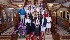 Participants at the June 2026 Policy Lab in Stresa pose for a "family photos" on a wooden staircase, with the ornate decor of the Hotel Regina surrounding them