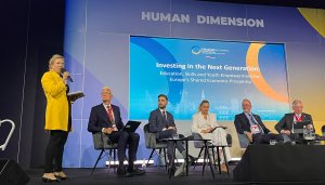 Dressed in yellow and off centre, ETF Director Pilvi Torsti stands upright, microphone in hand and addresses and audience, while other panel members remain seated, perched in front of a blue digital presentation scree, concentrate and listen