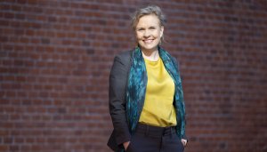 A blonde smiling woman, slightly off centre, wears a yellow satin blouse, a patterned dark teal scarf and grey blazer, with a brick wall occupying the background