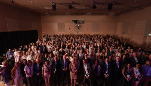 Facing the camera, a large number of attendees at the Forum on Vocational Excellence 2026, each wearing blue lanyards, pose for a picture in a large, wood-clad assembly hall