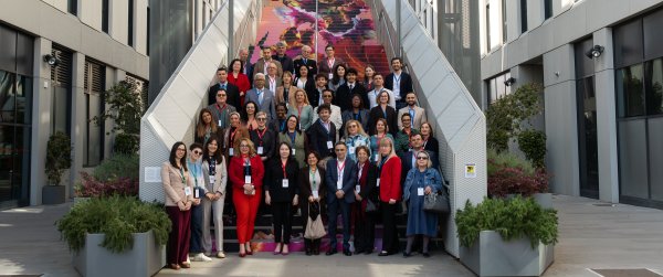 Various diversely clad experts and professionals stand for a group picture outdoors, at the foot of a grey, modern staircase, located between two offices 