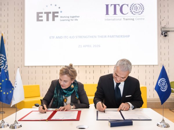 On a white table, a blonde agency director with a grey blazer and green scarf signs an agreement alongside a grey-haired gentleman wearing a suit