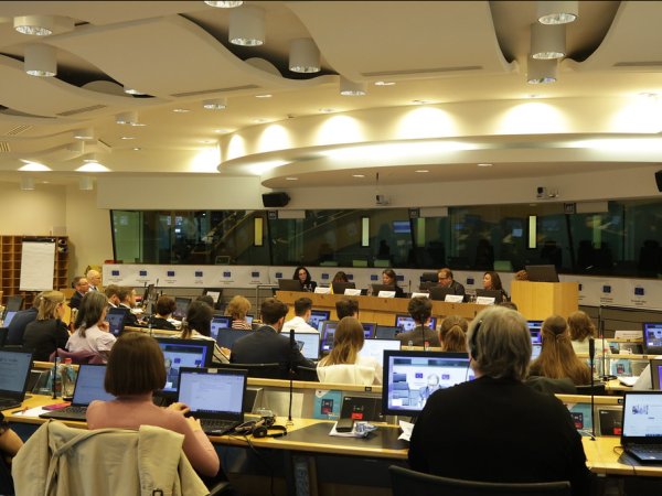 Various attendees seated in a semi-circular conference room, each equipped with laptops, listening to speakers address them from a multi-person central rostrum