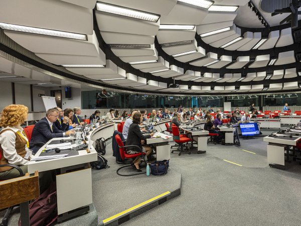 A brightly lit, largely white and grey hemicircular conference room, viewed in side profile, filled with people sitting down and observing the centre