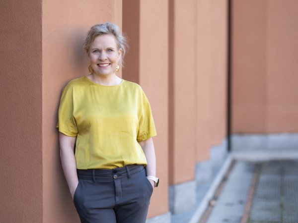 Pilvi Torsti, wearing a yellow satin top and grey slacks, smiling and leaning against the terracotta-coloured faced of a colonnade-ripple building