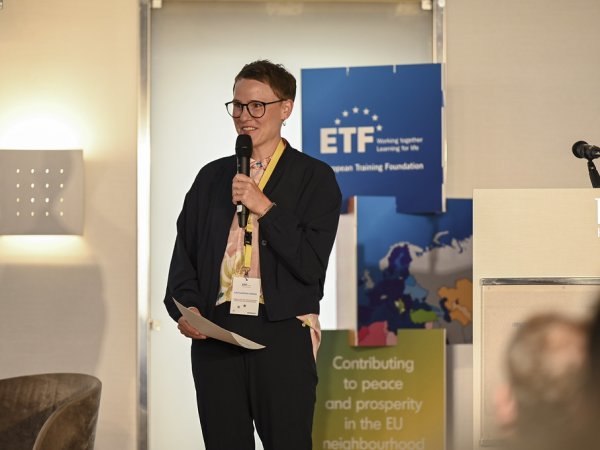 A short-haired young woman, piece of white paper in hand, wearing glasses, a grey jacket and a lanyard, holding a microphone as she addresses the audience from a stage, with a white lectern to her left and a blue ETF-branded totem located next to it