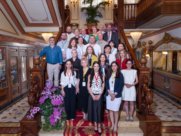 Participants at the June 2026 Policy Lab in Stresa pose for a "family photos" on a wooden staircase, with the ornate decor of the Hotel Regina surrounding them