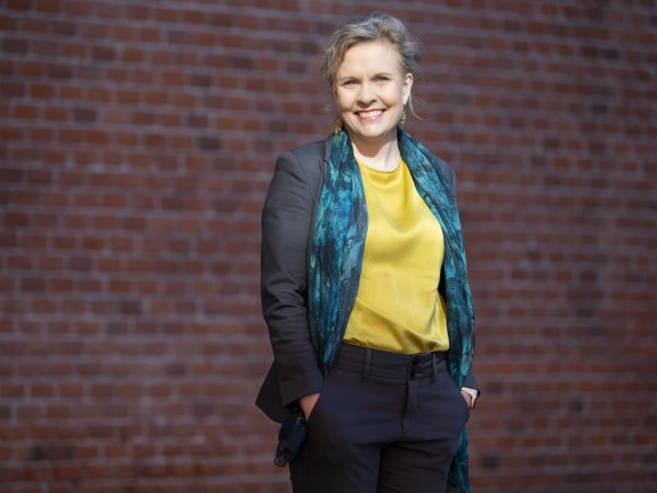 A blonde smiling woman, slightly off centre, wears a yellow satin blouse, a patterned dark teal scarf and grey blazer, with a brick wall occupying the background