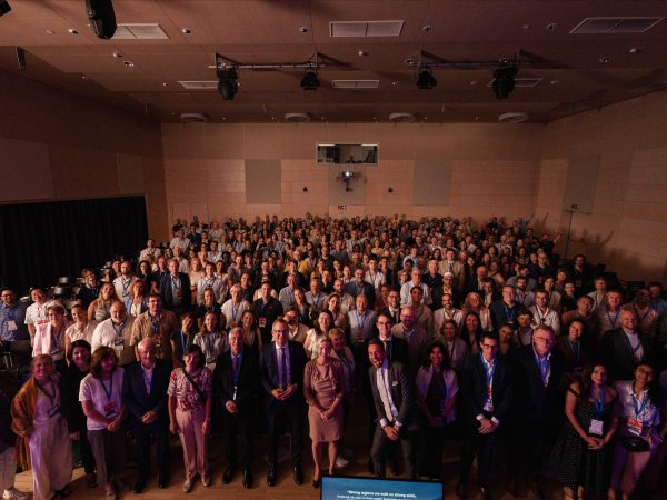 Facing the camera, a large number of attendees at the Forum on Vocational Excellence 2026, each wearing blue lanyards, pose for a picture in a large, wood-clad assembly hall