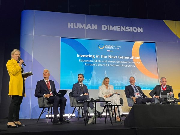Dressed in a yellow blazer and situated to the left of the frame, ETF Director Pilvi Torsti addresses a conference room, microphone in hand, with other panellists sitting to her right watching her deliver her address