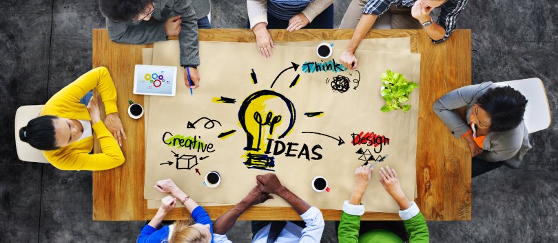 group of people (seen from above) sitting around a table and jointly creating a design, 'Ideas' being the central theme