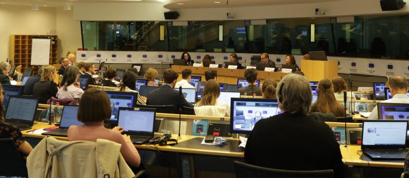 Various attendees seated in a semi-circular conference room, each equipped with laptops, listening to speakers address them from a multi-person central rostrum