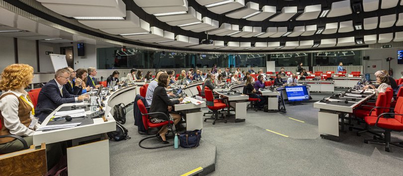 A brightly lit, largely white and grey hemicircular conference room, viewed in side profile, filled with people sitting down and observing the centre