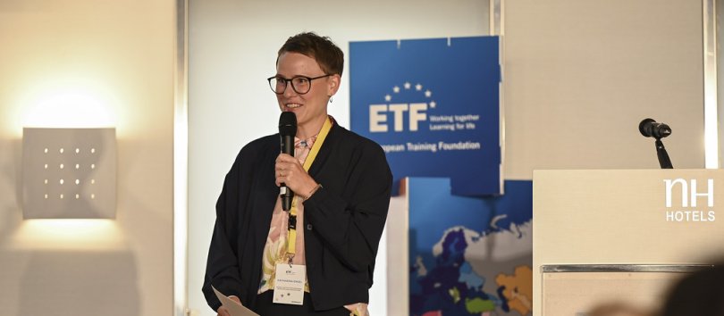 A short-haired young woman, piece of white paper in hand, wearing glasses, a grey jacket and a lanyard, holding a microphone as she addresses the audience from a stage, with a white lectern to her left and a blue ETF-branded totem located next to it