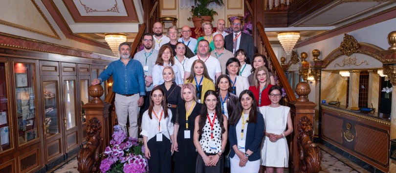 Participants at the June 2026 Policy Lab in Stresa pose for a "family photos" on a wooden staircase, with the ornate decor of the Hotel Regina surrounding them