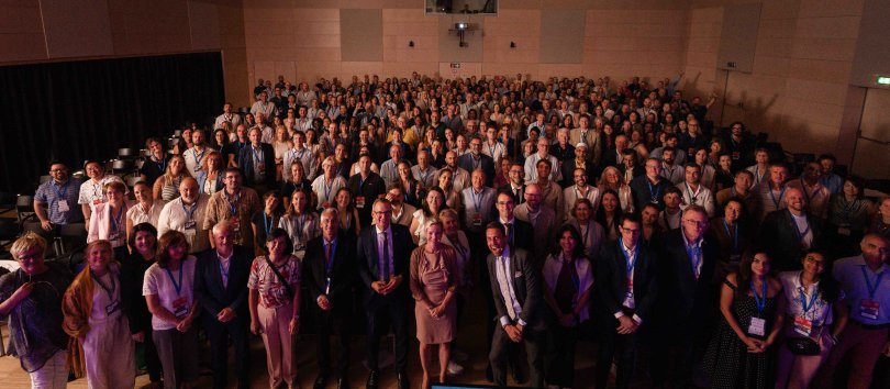 Facing the camera, a large number of attendees at the Forum on Vocational Excellence 2026, each wearing blue lanyards, pose for a picture in a large, wood-clad assembly hall