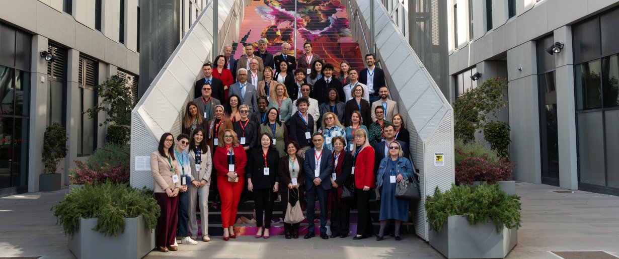 Various diversely clad experts and professionals stand for a group picture outdoors, at the foot of a grey, modern staircase, located between two offices 