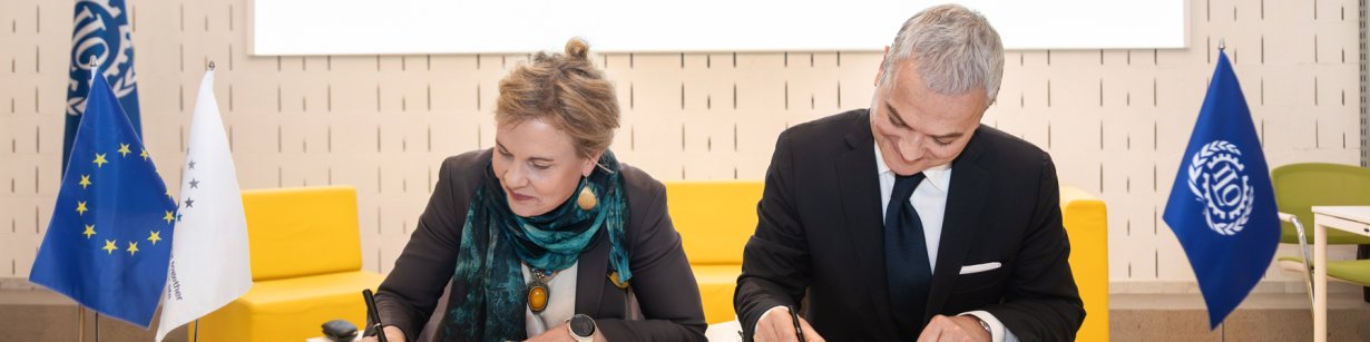 On a white table, a blonde agency director with a grey blazer and green scarf signs an agreement alongside a grey-haired gentleman wearing a suit