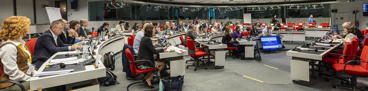 A brightly lit, largely white and grey hemicircular conference room, viewed in side profile, filled with people sitting down and observing the centre