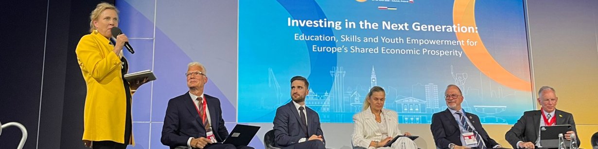 Dressed in yellow and off centre, ETF Director Pilvi Torsti stands upright, microphone in hand and addresses and audience, while other panel members remain seated, perched in front of a blue digital presentation scree, concentrate and listen