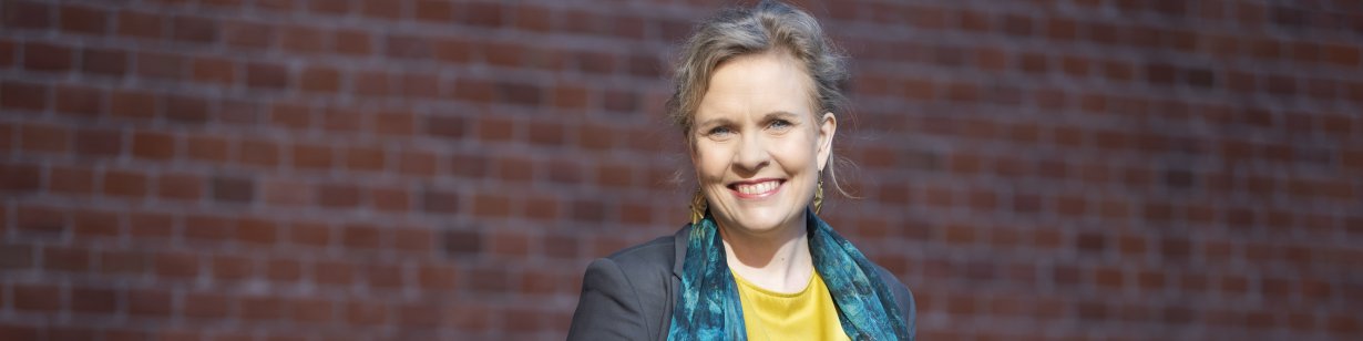 A blonde smiling woman, slightly off centre, wears a yellow satin blouse, a patterned dark teal scarf and grey blazer, with a brick wall occupying the background